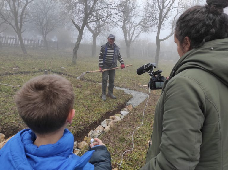 a worker building a cottage foundation and being filmed by a woman and a boy